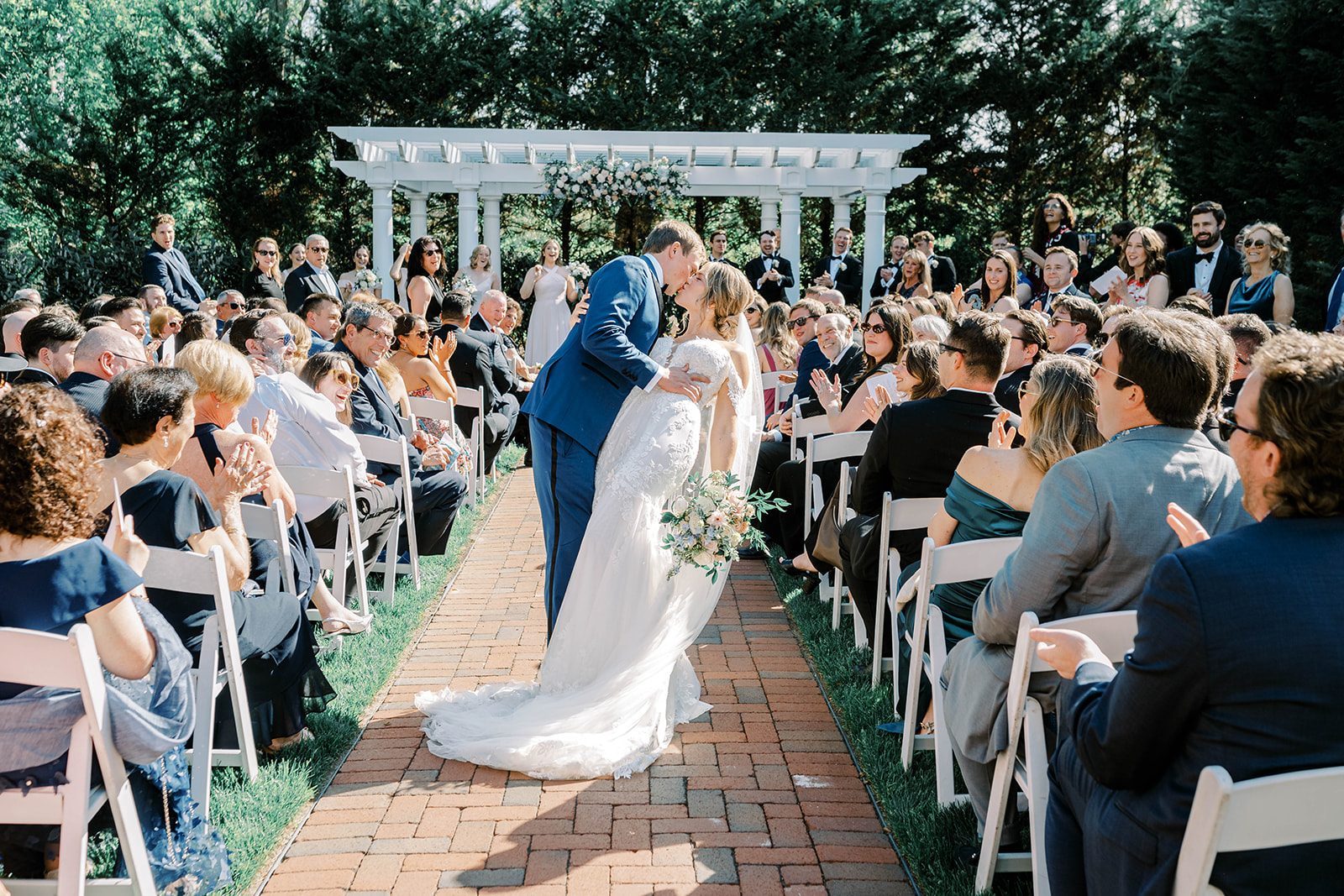 Newlyweds kiss in the brick aisle as guests applaud before the garden pergola